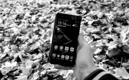 Black and white image of a hand holding a smartphone displaying social media apps, surrounded by fallen leaves.